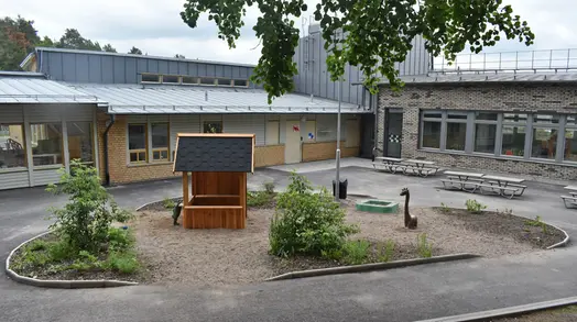 Utegård på en förskola. På en asfalterad yta framför en tegelbyggnad finns en lekyta med ett offentligt konstverk. Verket består av ett lekhus i trä och två bronsfigurer.