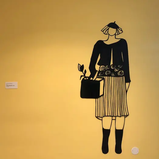 En gul vägg med ett svart konstverk, en relief i lackad plåt. Verket föreställer en tecknad kvinna i kjol och stövlar, med en väska på armen och en basker på huvudet.